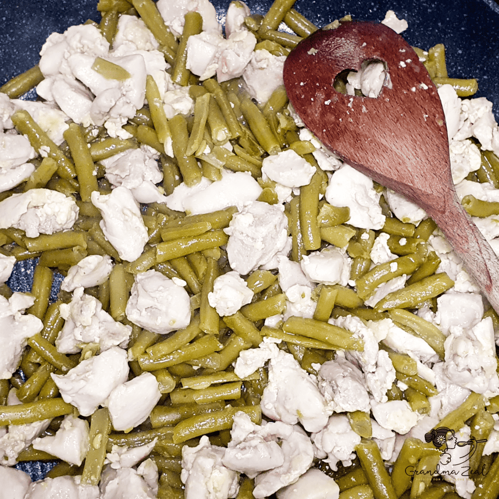 Green beans and sliced chicken cooking together in a pan, stirred with a wooden spoon.
