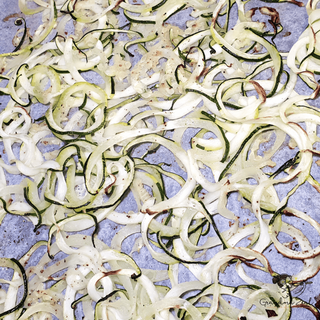 Baked courgette zoodles on a parchment-lined tray, slightly wilted and golden around the edges.