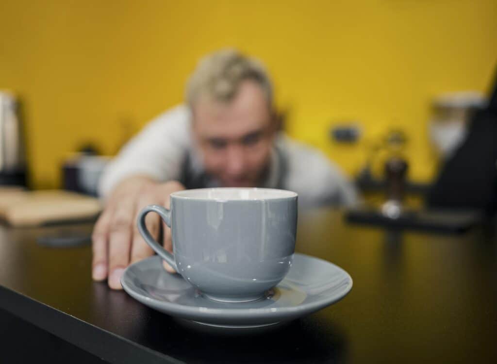 Cup of coffee in the foreground with a blurry man reaching for it — a visual cue for fatigue and reliance on caffeine