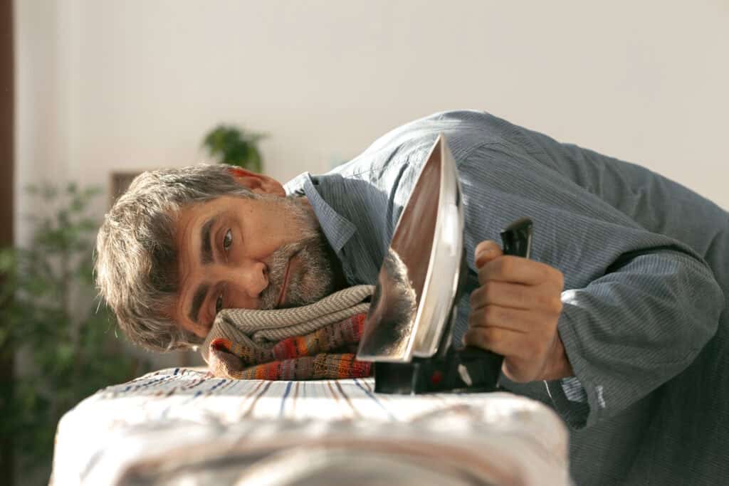 Tired man holding an iron, head resting  on a stack of freshly ironed clothes featured morning fatigue
