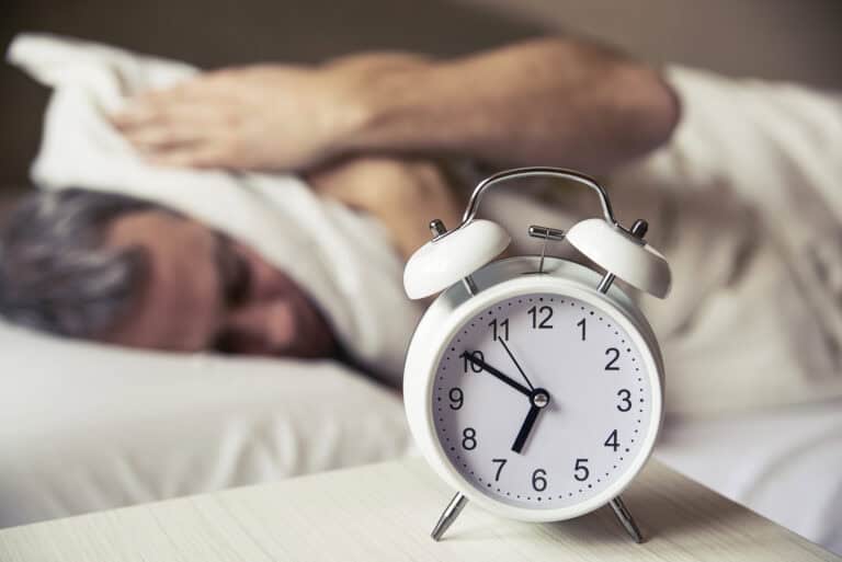 Sleepy man covering ears with pillow as he looks at alarm
