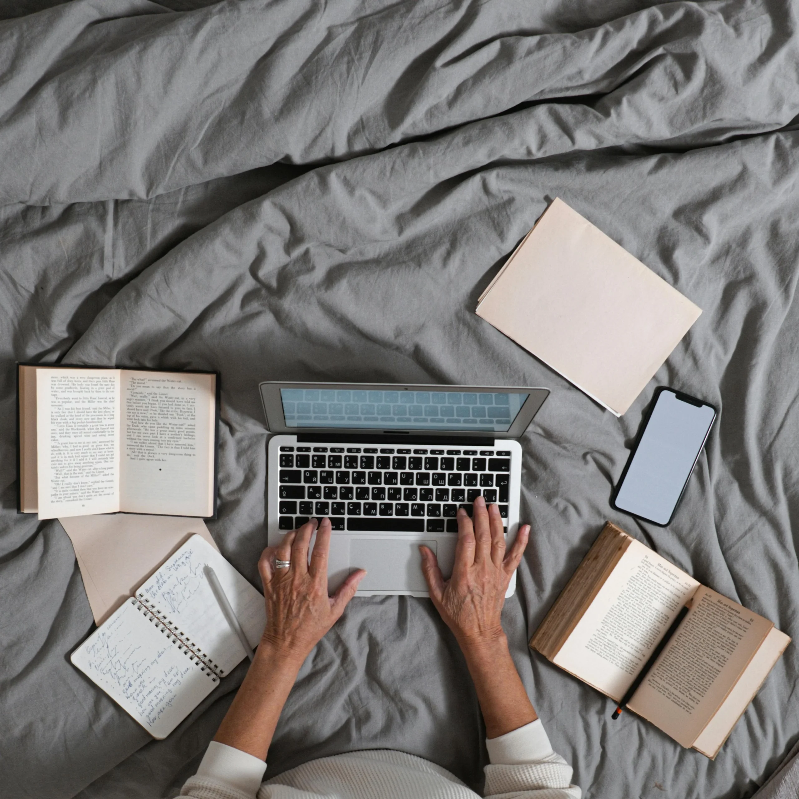Hands typing on a laptop placed on a bed, surrounded by books and notebooks; creating, learning, and writing