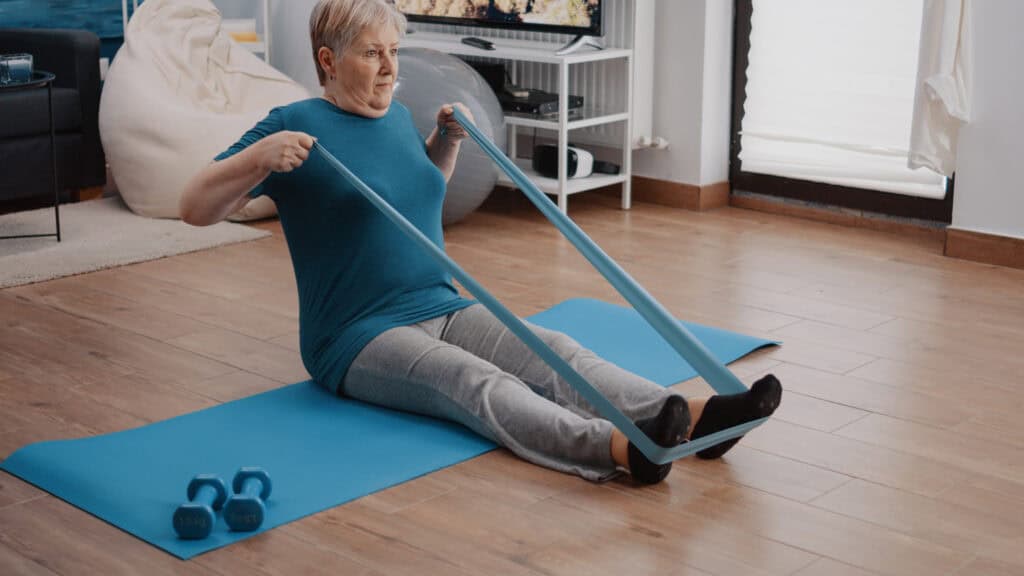 Senior woman pulling flexible belt to stretch arms and legs, doing strength training with sport equipment preventing osteoporosis through exercise