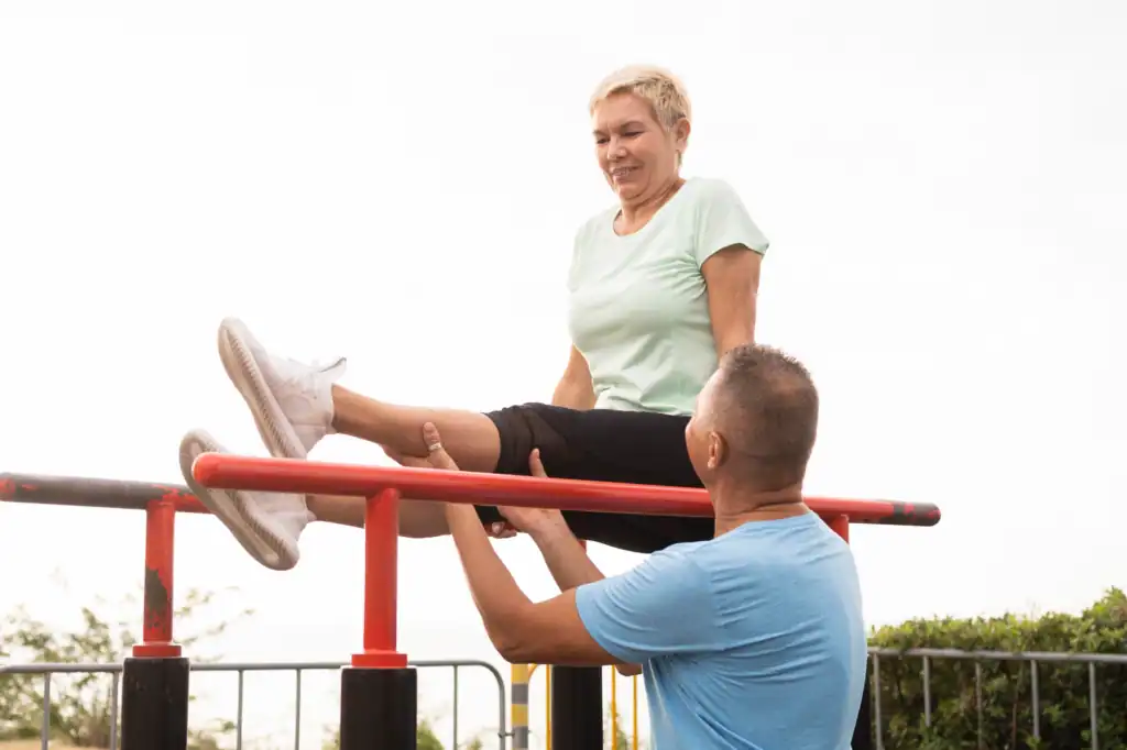 women in her 50s exercising on the parallel bars outdoor with the help of the instructor