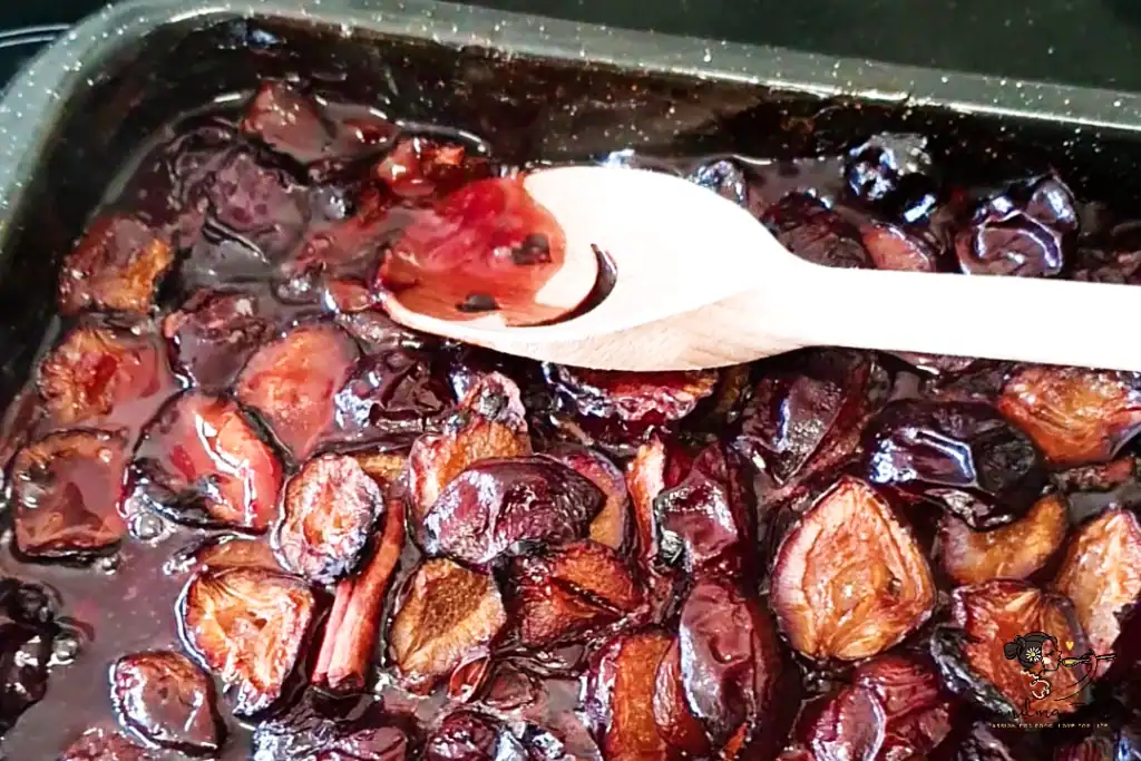 Partially baked plums in a tray after one hour, with a wooden spoon pressing the top layer down into the juices