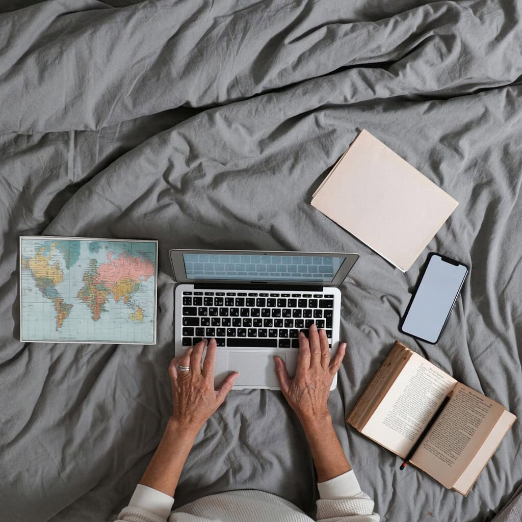Hands typing on a laptop placed on a bed, surrounded by book, notebook and map creating, learning, and writing