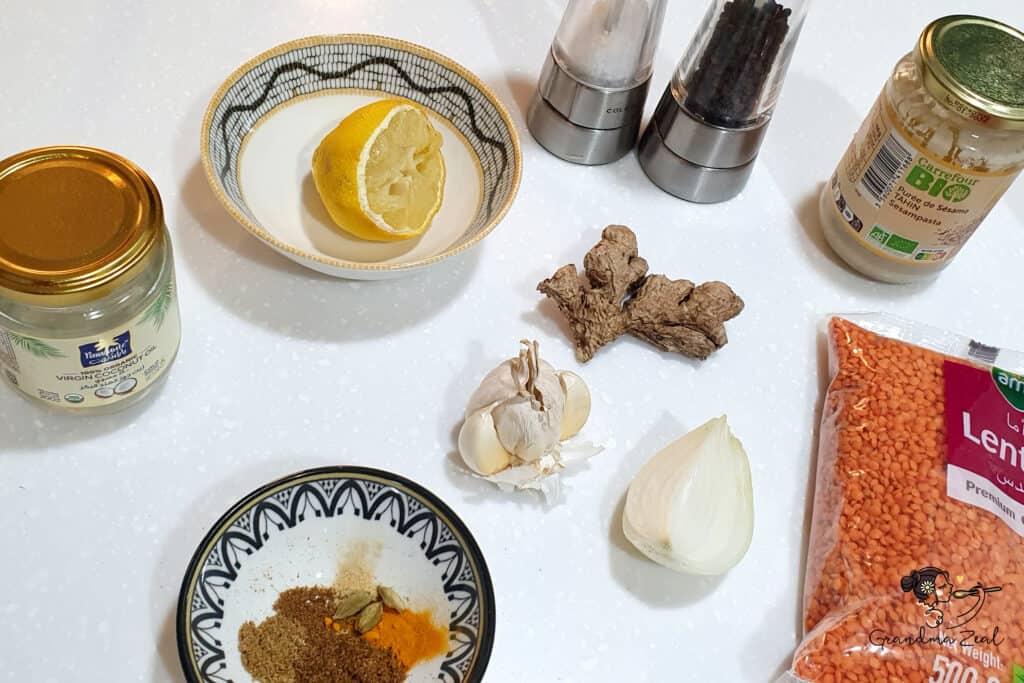 Ingredients for a lentil hummus recipe laid out on a white counter, including red lentils, lemon, garlic, ginger, onion, spices, coconut oil, tahini, salt and pepper.