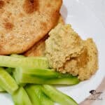 A white plate with creamy lentil humus, sliced cucumber sticks, and two pieces of seasoned flatbread.