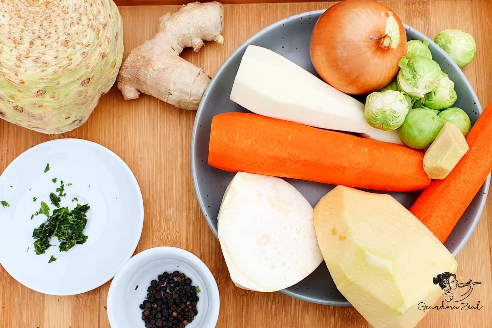 Fresh soup vegetables arranged on a wooden board, including carrots, celeriac, parsnip, onion, Brussels sprouts, ginger, black peppercorns, and chopped lovage.